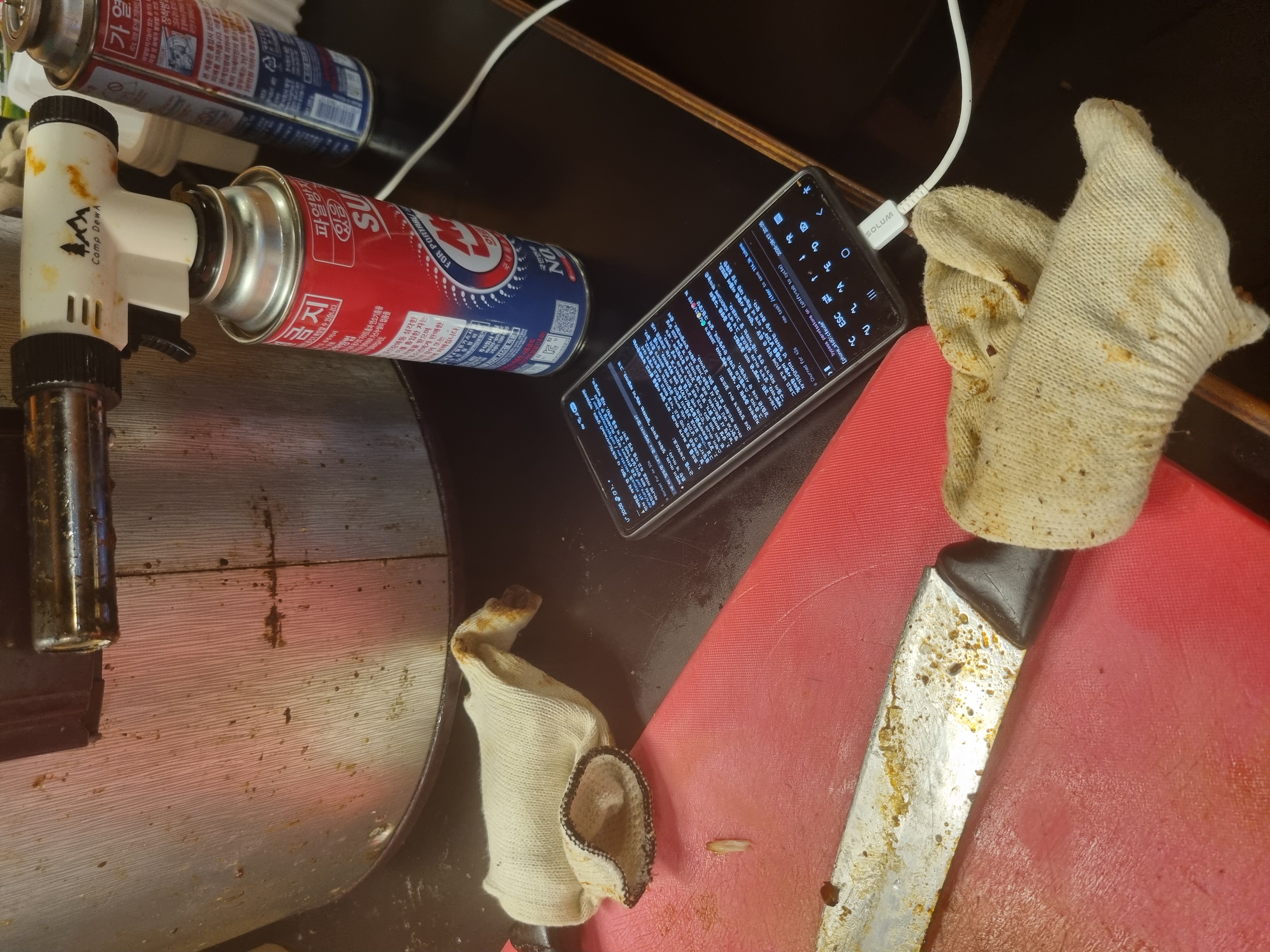 The real worksite — grease-stained pots, soiled gloves, a knife and a torch, and in the middle, a phone with a terminal open